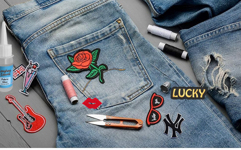 A pair of blue jeans decorated with various patches including a rose, guitar, and lips, alongside sewing supplies like scissors, thread, and fabric glue on a table.