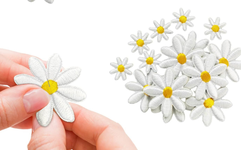 Hand holding an embroidered white daisy with a yellow center, alongside a scattered pile of similar daisies on the right.