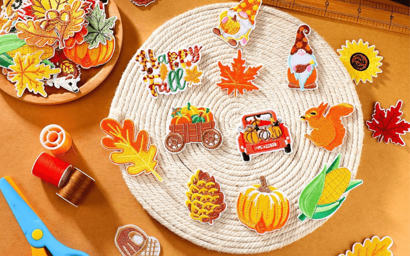 A collection of colorful fall-themed embroidered patches arranged on a woven mat, including leaves, pumpkins, a squirrel, and a red truck. Scissors, threads, and a ruler are nearby.
