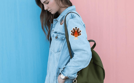 Young woman wearing a denim jacket with a turkey patch on the sleeve and carrying a green backpack, stands against a split light blue and pink background.
