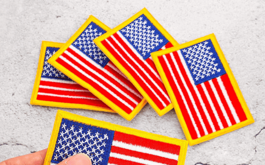 A hand holds one of several USA flag patches, each outlined in yellow, against a gray textured background.
