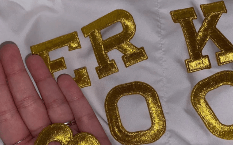 Close-up of a hand touching gold letters "erk" on a white fabric surface.
