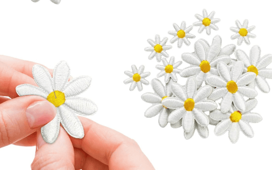 Hand holding an embroidered white daisy with a yellow center, alongside a scattered pile of similar daisies on the right.
