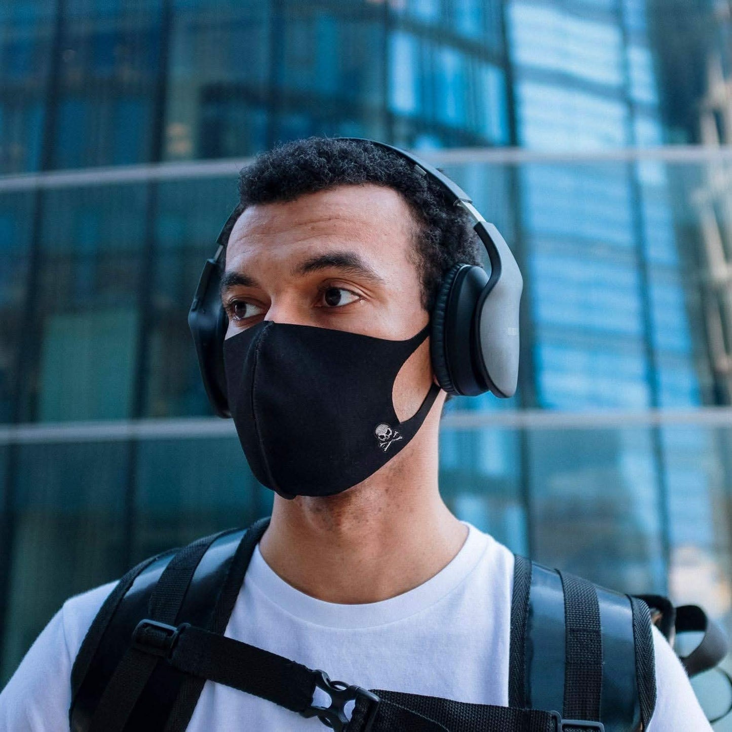 A person wearing headphones, a black face mask, and a white shirt stands in front of a glass building with a backpack featuring a Laughing Lizards Extra Small Skull Halloween Embroidered Iron On Patch.