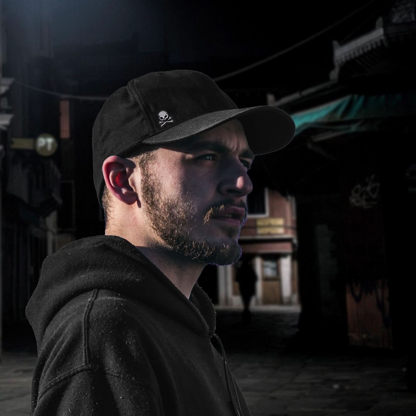A man in a black hoodie and cap stands in a dimly lit urban street, looking serious. His outfit is accented by Laughing Lizards' Extra Small Skull Halloween Embroidered Iron On Patch (10 Pack).