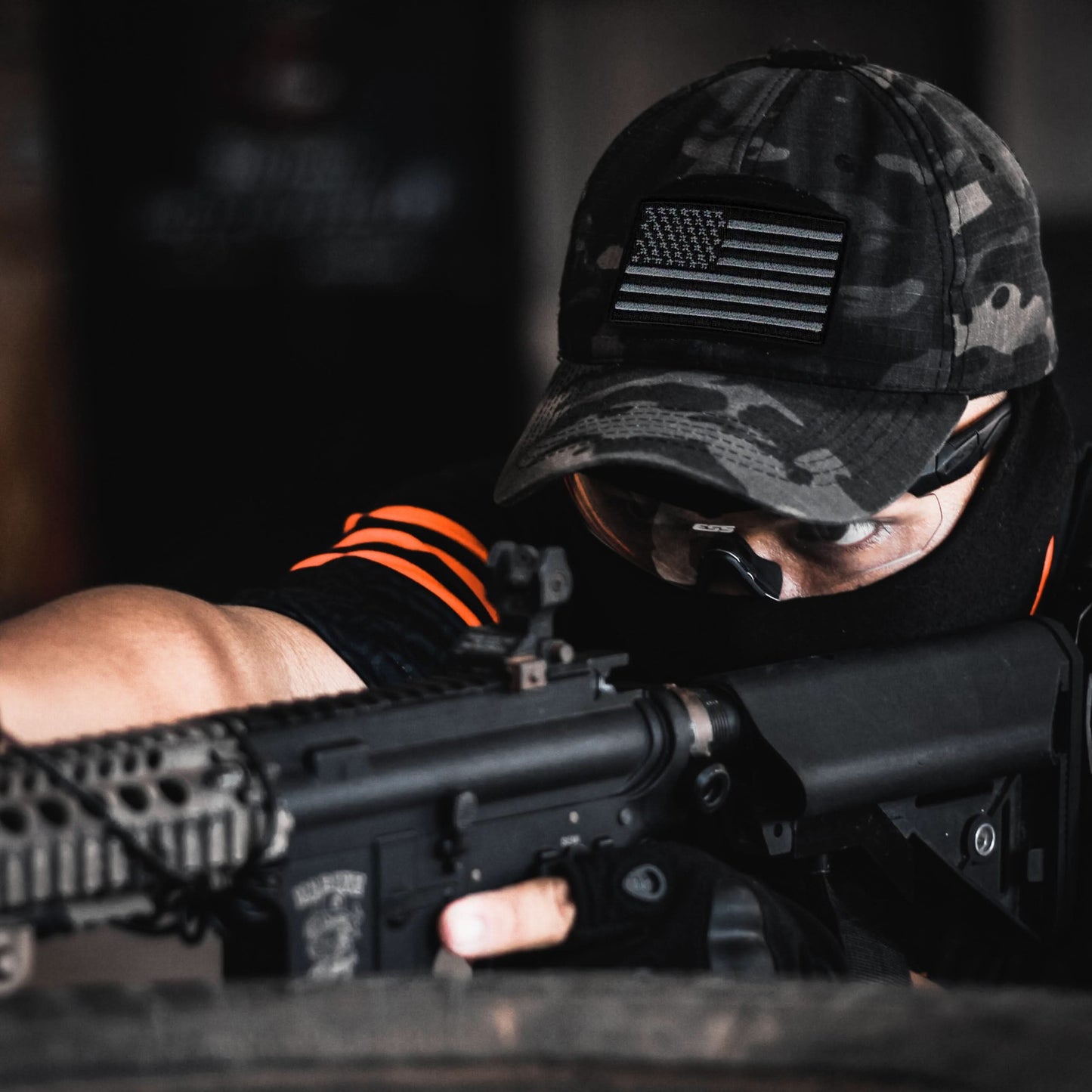 A person in tactical gear and a camouflage cap with a Laughing Lizards Black & Gray USA Flag Embroidered Patch aims a rifle, the scope and barrel partially obscuring their face.