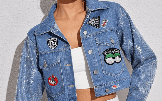 Person wearing a blue denim jacket with sequin sleeves and various colorful patches, over a white crop top, standing against a light-colored wall.