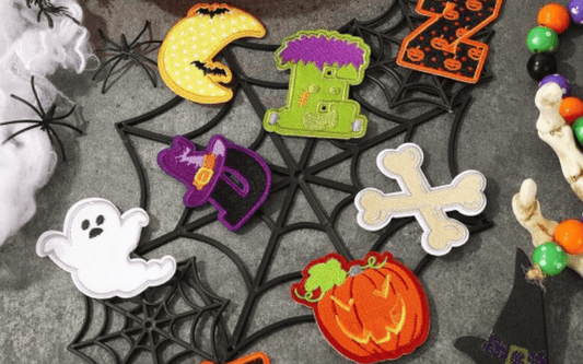 A Halloween-themed assortment of patches, including a ghost, pumpkin, moon, and Frankenstein's head, displayed on a spiderweb mat. Nearby are plastic spiders, beads, and a witch hat.