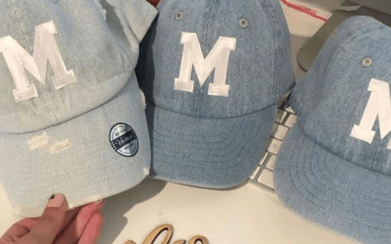 Three light blue denim caps with the letter "M" embroidered in white are placed on a white desk. One cap has a manufacturer's sticker on the brim.
