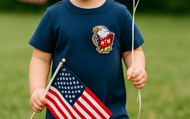 A child wearing a blue shirt with an eagle patch holds a small American flag and white balloons, standing outdoors on grass.