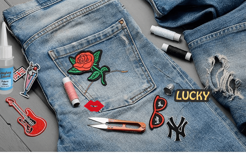 A pair of blue jeans decorated with various patches including a rose, guitar, and lips, alongside sewing supplies like scissors, thread, and fabric glue on a table.