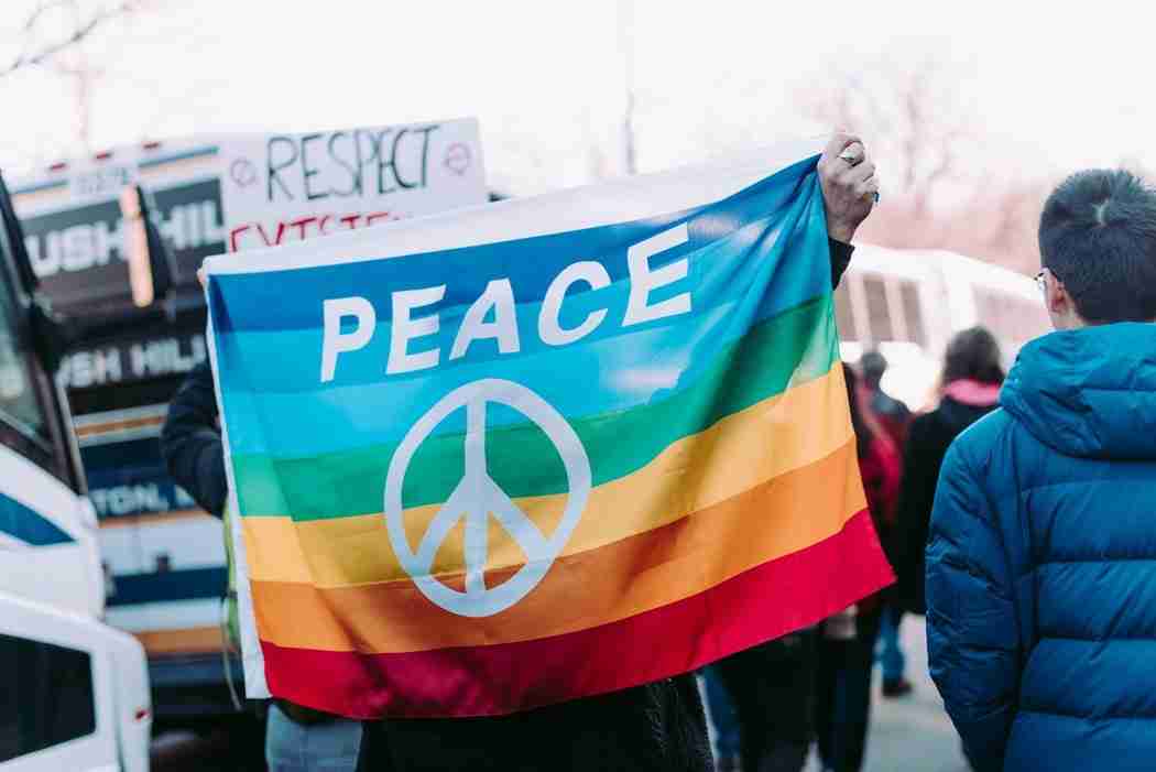 Person holds a rainbow-colored flag with the word "PEACE" and a peace symbol at a public gathering; people and buses are visible in the background.
