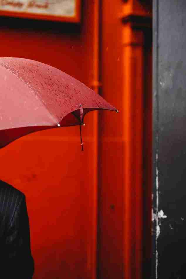 A red umbrella with raindrops on it is partially open against a bright red wall and a dark vertical structure on the right.