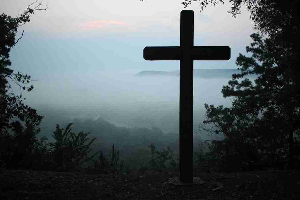 A large wooden cross stands on a hilltop surrounded by trees, overlooking a foggy valley at dawn or dusk.
