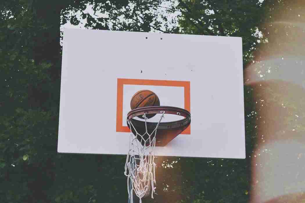 A basketball rests on the rim of an outdoor basketball hoop with a white backboard, surrounded by trees in the background.