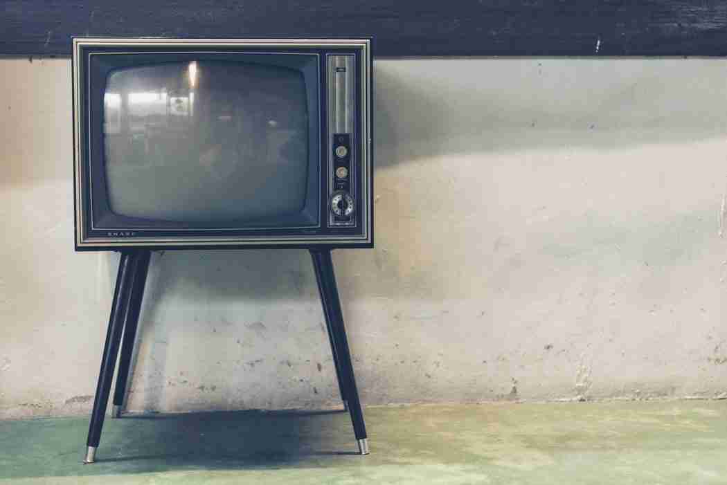A vintage television set with antennas stands on four legs against a plain wall and green-tinted floor.