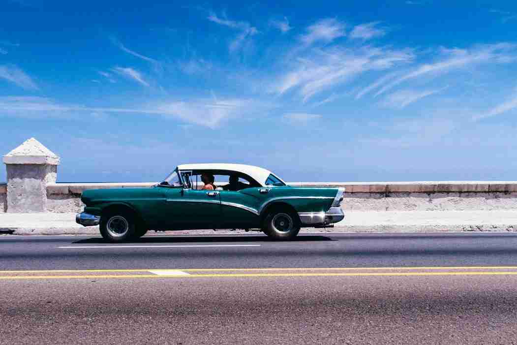 A vintage green and white car is parked on a road beside a stone wall with the ocean and a blue sky in the background.