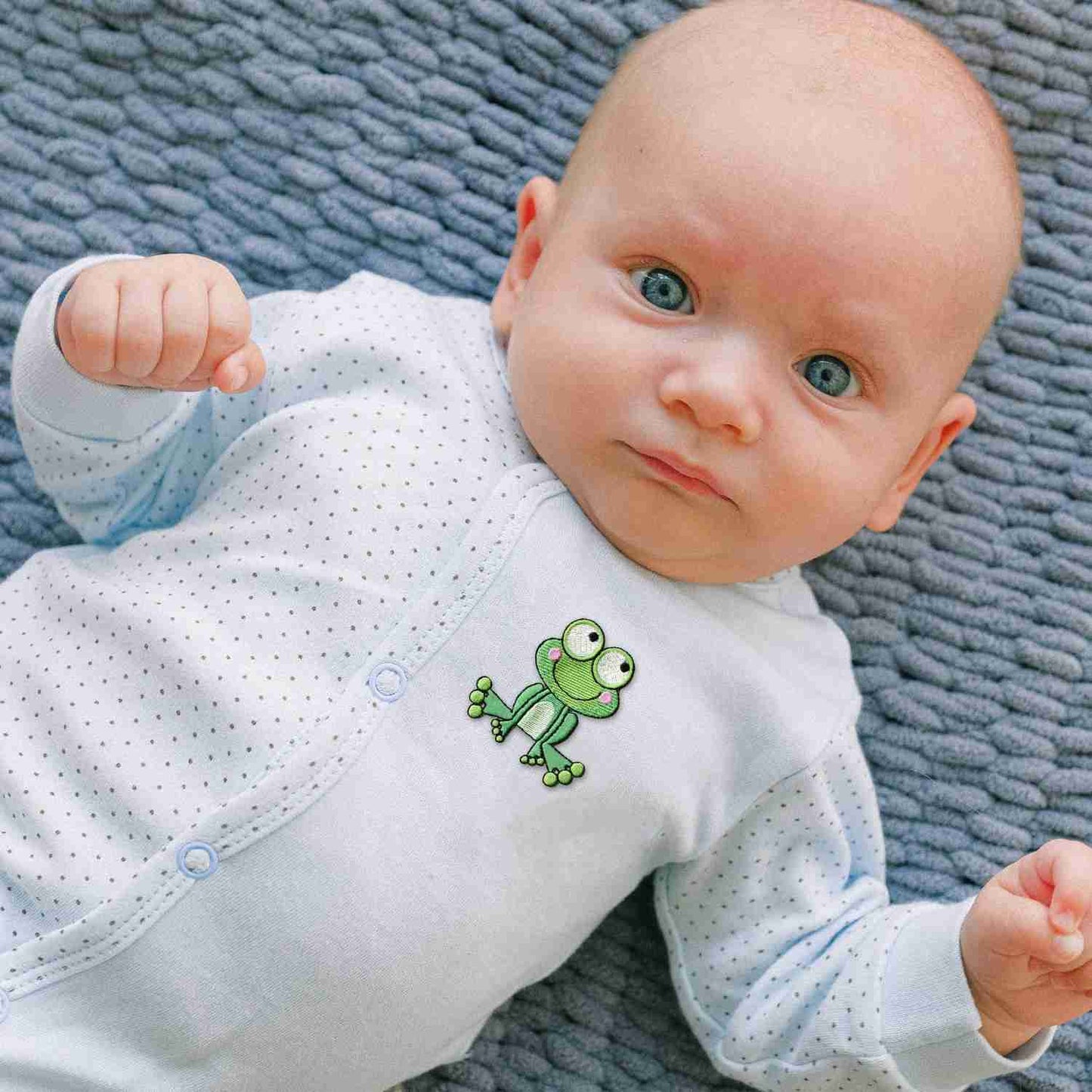 A baby laying on a blue blanket with Frog Patches (2 Pack) Animal Embroidered Iron on Patch Appliques on it.