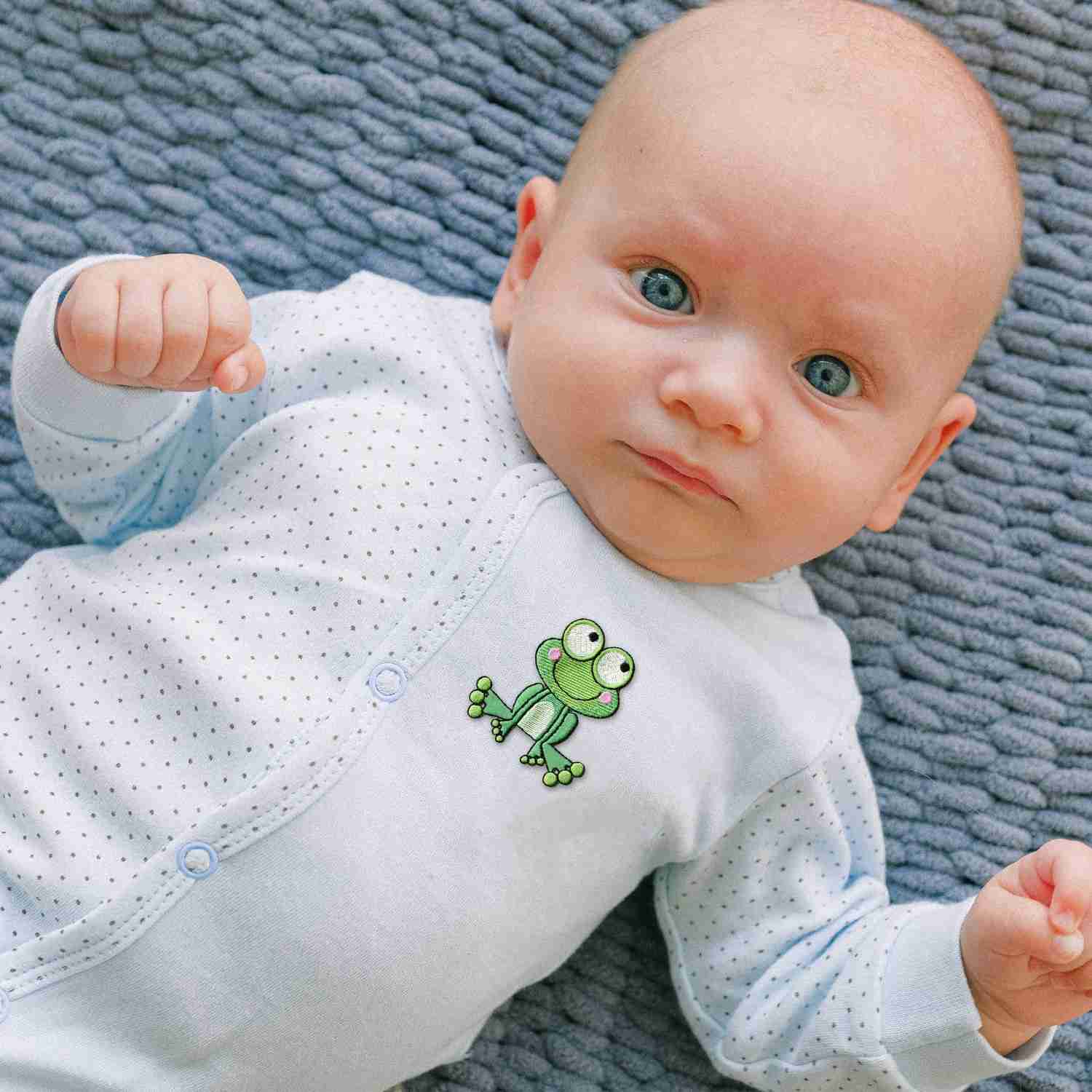 A baby laying on a blue blanket with Frog Patches (2 Pack) Animal Embroidered Iron on Patch Appliques on it.