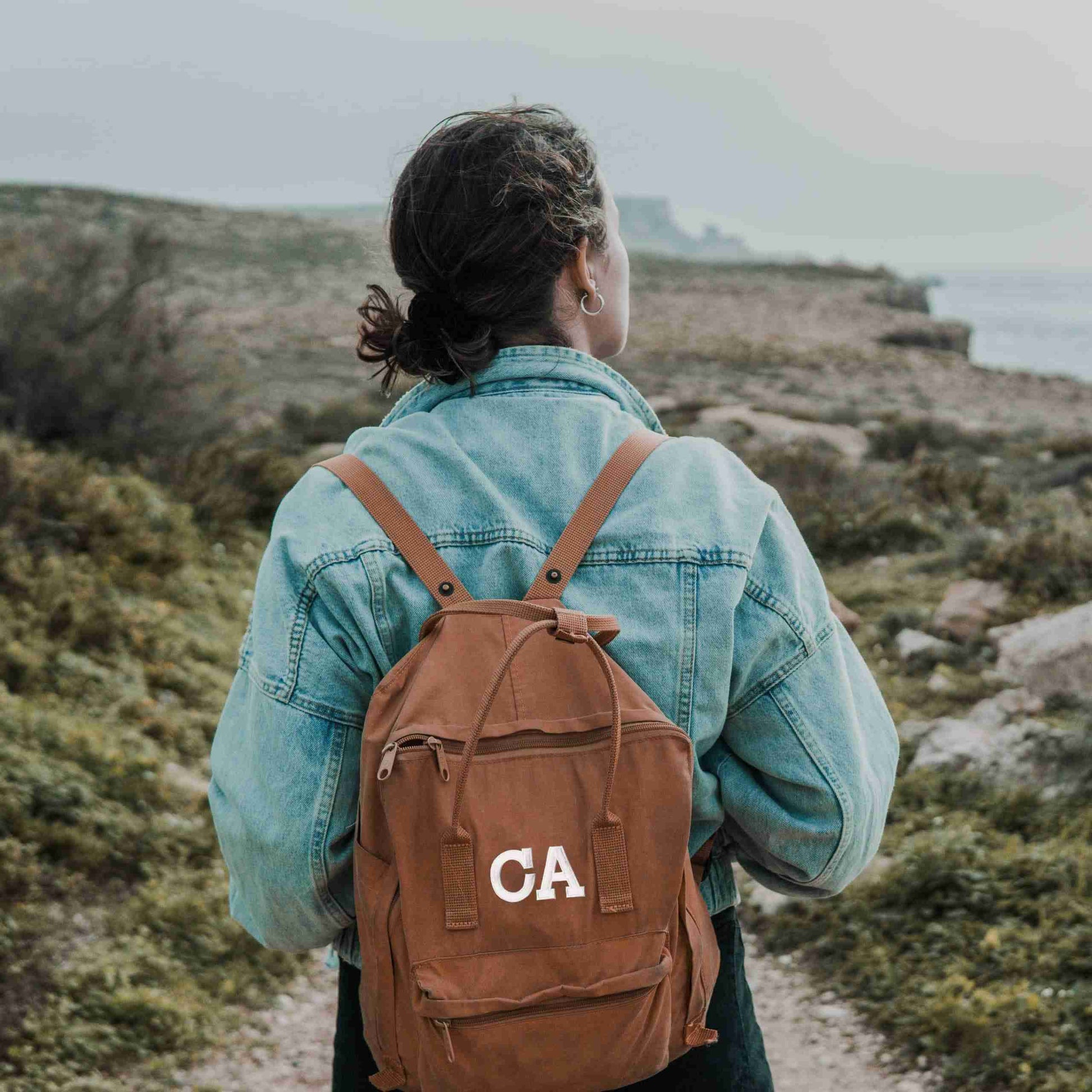 A woman wearing a brown backpack with the Iron On Letter Patches, 2" Block Letters & Numbers on it.
