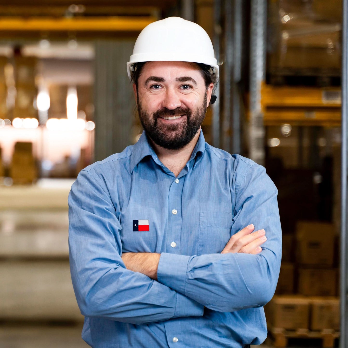 A bearded man in a white hard hat and blue shirt stands with arms crossed in a warehouse, proudly showing off the Laughing Lizards Small Texas Flag Patch from the 10-pack embroidered iron-on applique on his sleeve.