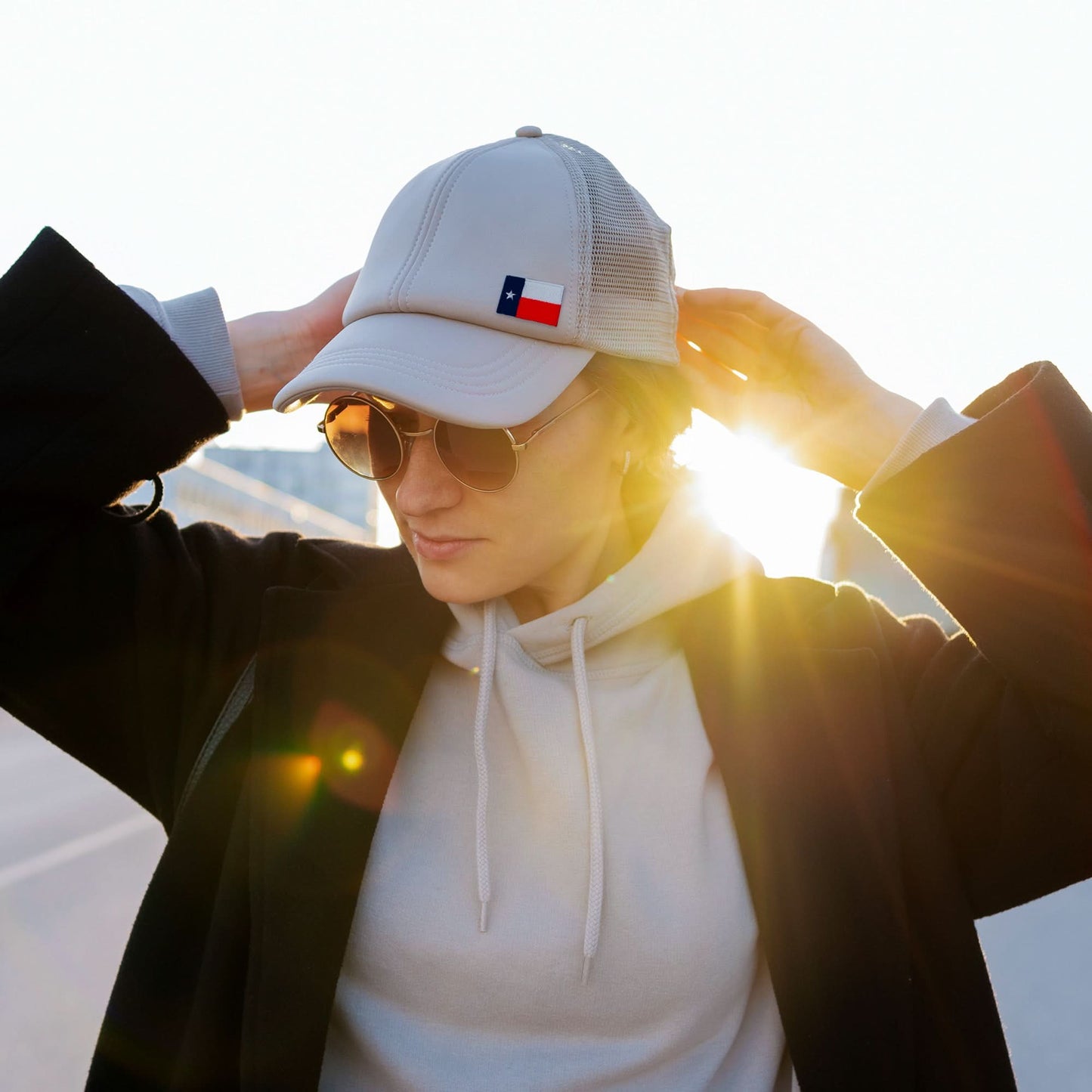 Person wears sunglasses, a light hoodie, and a cap with a Laughing Lizards Small Texas Flag Patch from the 10-Pack embroidered applique set, adjusting their hat outdoors in the sunlight.